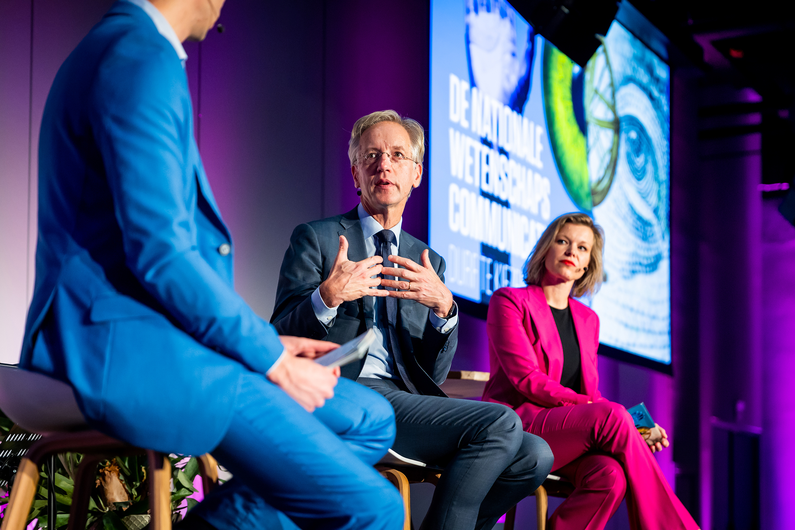 Drie mensen zitten op het podium onder paarse verlichting, verwikkeld in een discussie. Event branding is zichtbaar op het grote scherm met Nederlandse tekst achter hen. De man in het blauwe pak houdt een kaart vast terwijl de anderen naar het publiek kijken en aandachtig luisteren.