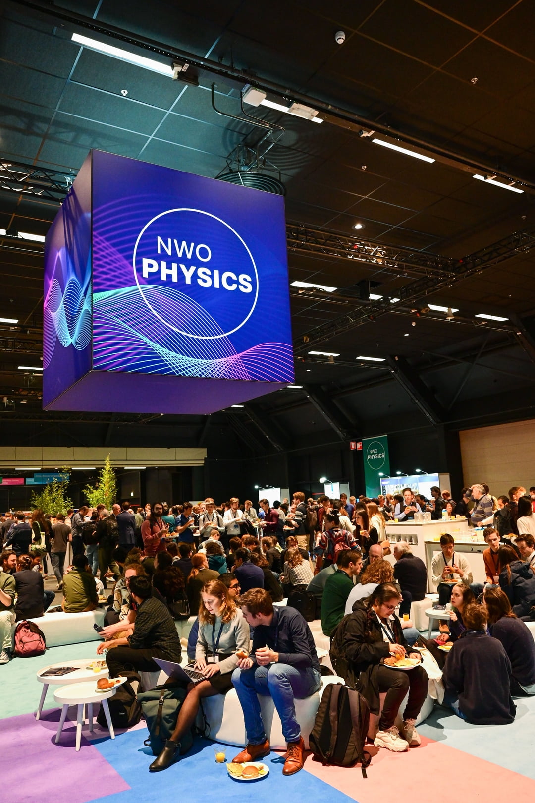 Een grote menigte verzamelt zich in een helder verlichte conferentiezaal onder een hangende kubus met het opvallende evenementlogo "NWO PHYSICS" met kleurrijke golven. Deelnemers socialiseren, eten en netwerken rond tafels en banken in een levendige sfeer.