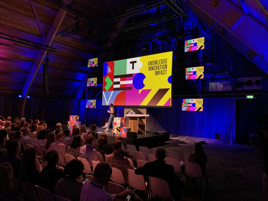 Een spreker staat op het podium voor een scherm met kleurrijke geometrische vormen en de woorden KNOWLEDGE INNOVATION IMPACT tijdens een conferentie. Deelnemers zitten met hun gezicht naar het podium in een modern auditorium met houten plafonds en paarse verlichting.